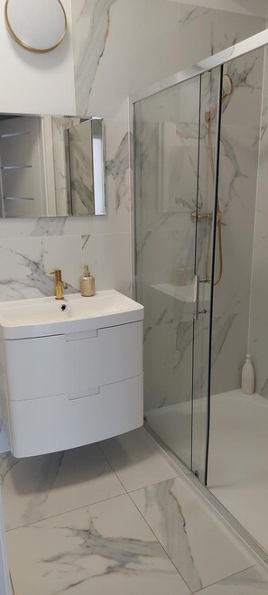 Un baño con suelo de mármol, tocador blanco con grifo dorado y zona de ducha con mampara de vidrio.