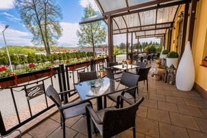 Un patio exterior con muebles de mimbre, una mesa de vidrio y vistas a un área ajardinada con árboles y flores.