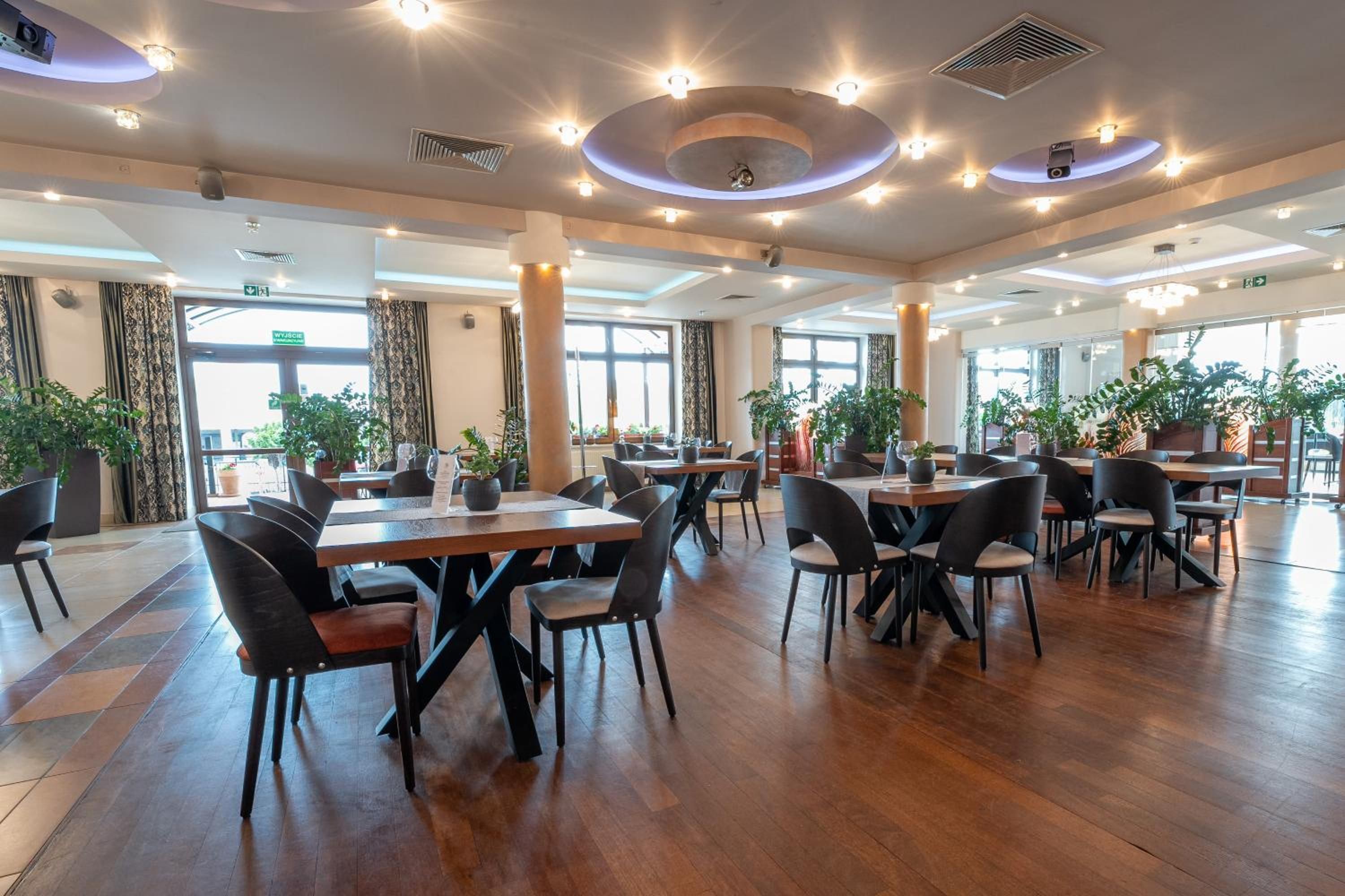 A spacious indoor dining area with multiple tables and chairs, large windows, and potted plants.