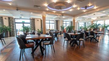 A spacious indoor dining area with multiple tables and chairs, large windows, and potted plants.