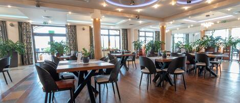 A spacious indoor dining area with multiple tables and chairs, large windows, and potted plants.
