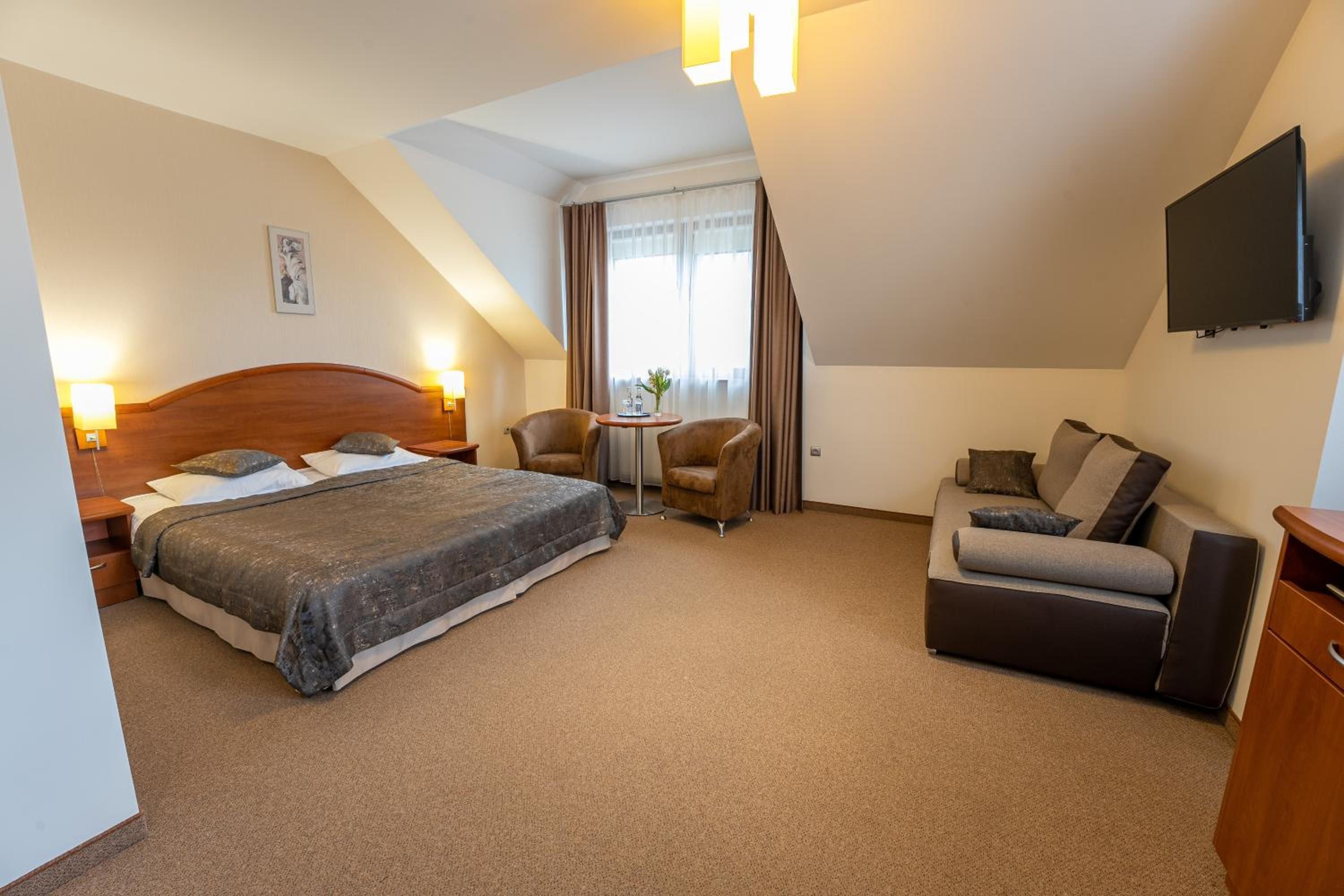 A hotel room with a bed, a sofa, a small table with chairs, a TV mounted on the wall, and a window with curtains.