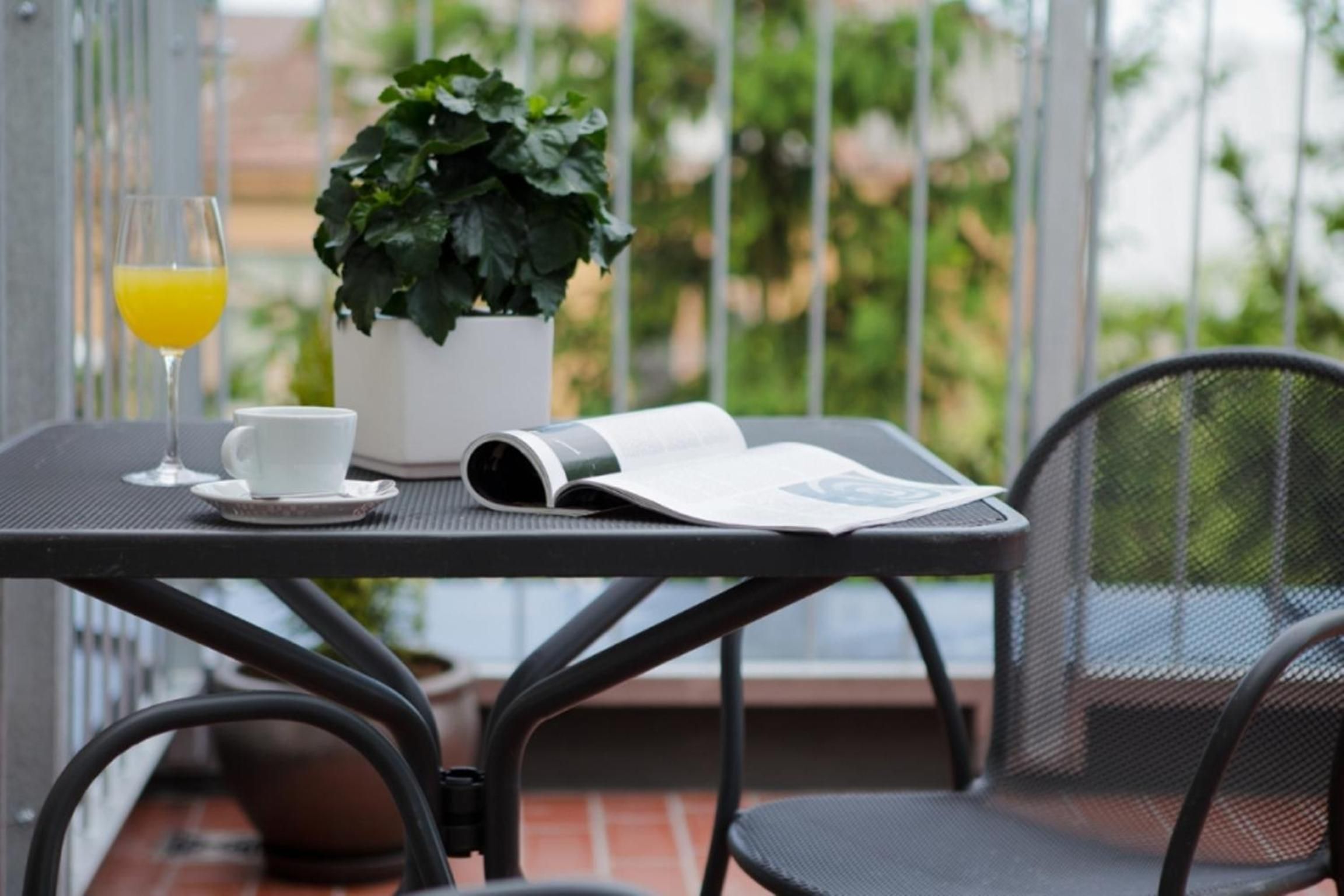 Una mesita con una planta en maceta, un vaso de zumo de naranja, una taza y una revista en un balcón.