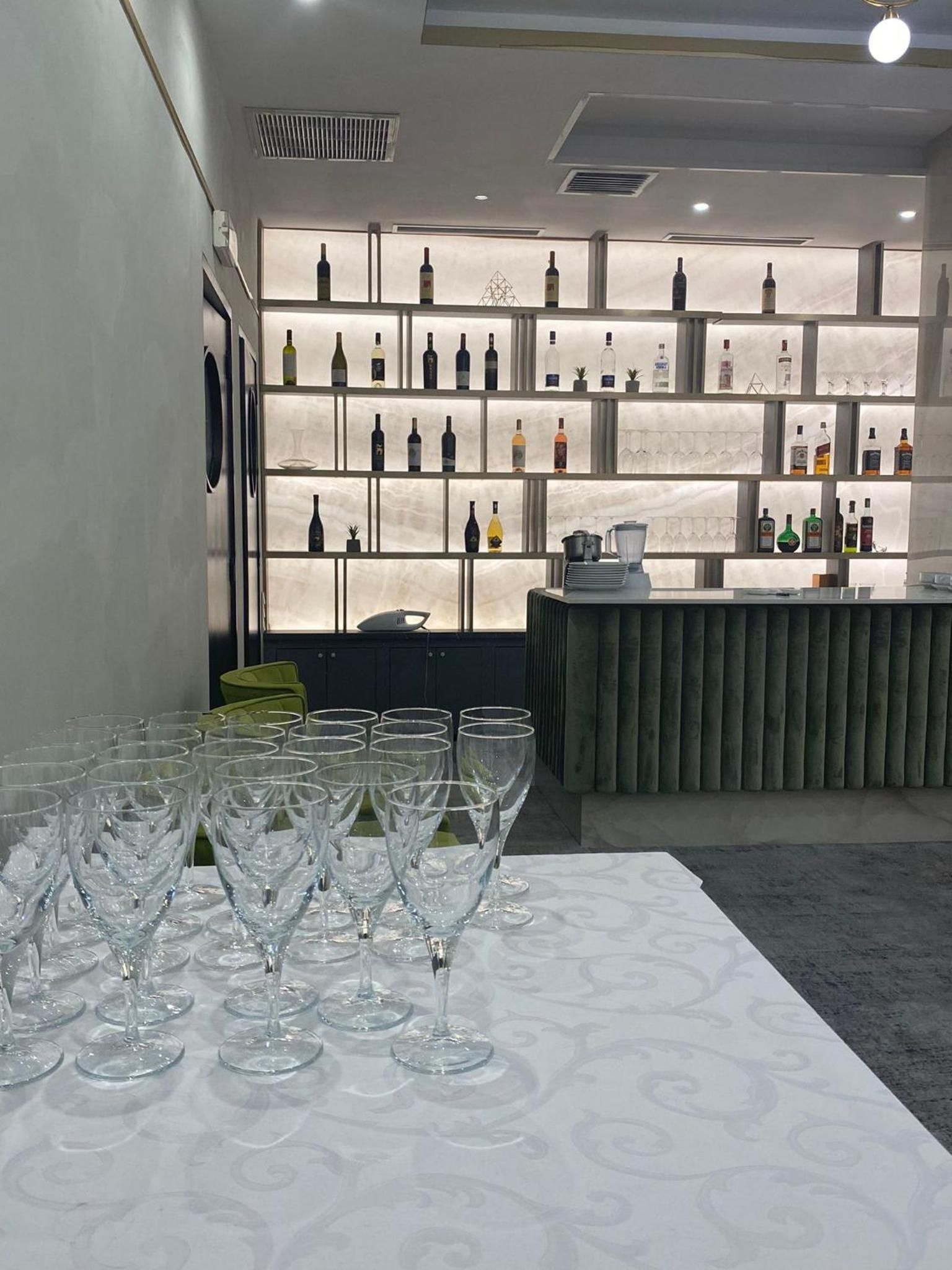 A bar area with a long table of empty wine glasses, a bar counter with a few items, and a wall of shelves displaying various bottles.