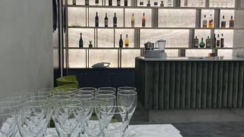 A bar area with a long table of empty wine glasses, a bar counter with a few items, and a wall of shelves displaying various bottles.