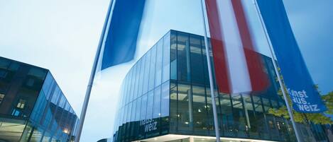 Un edificio moderno con fachada de vidrio y varias banderas, incluyendo una destacada con diseño azul, blanco y rojo.