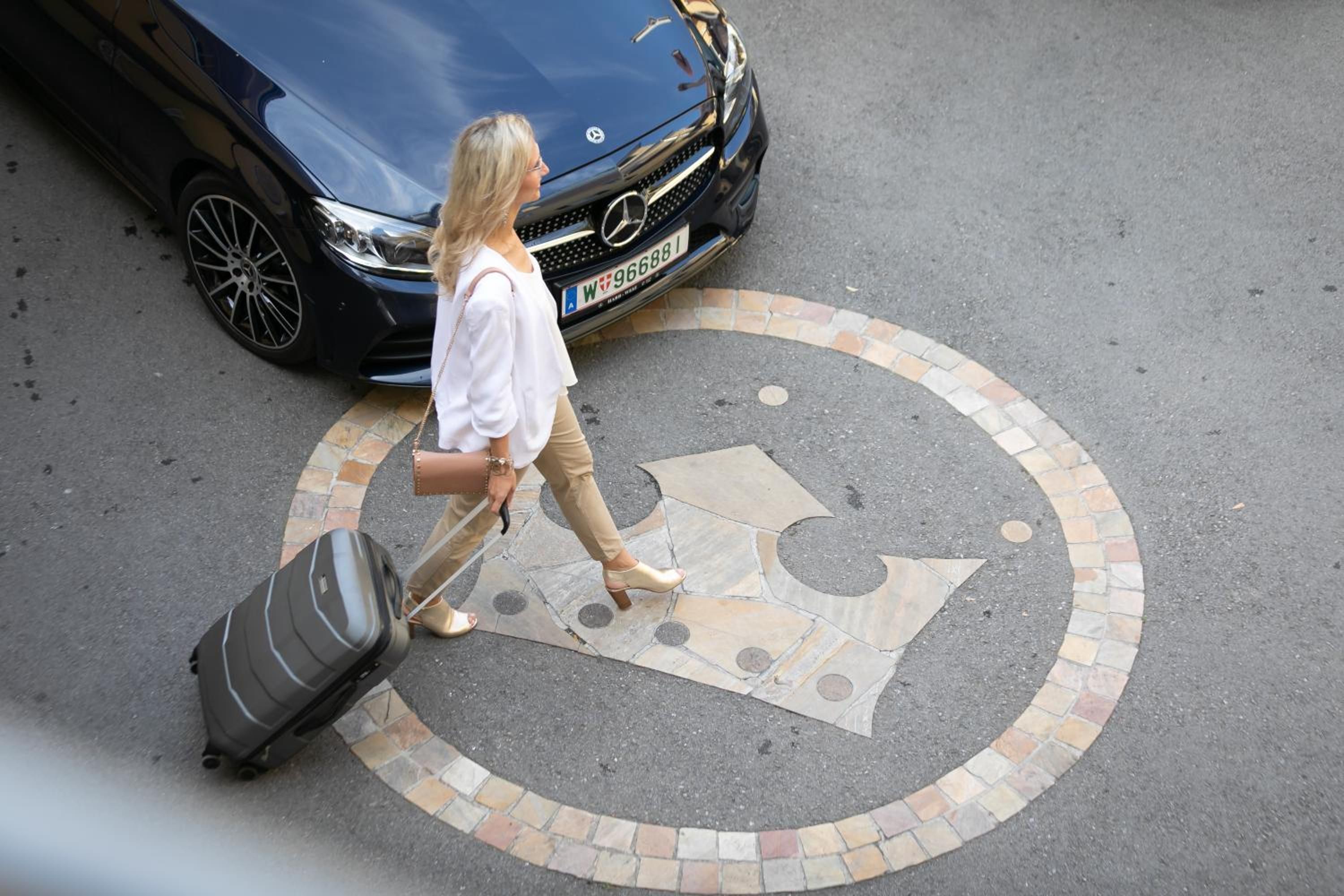 A person walking with a suitcase on a paved area with a car in the background.