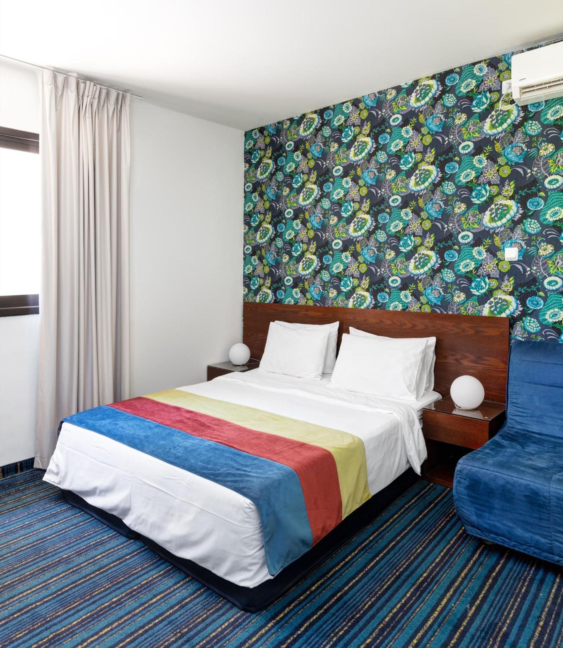 A hotel room with a bed, a blue armchair, a patterned wallpaper, and a window with curtains.