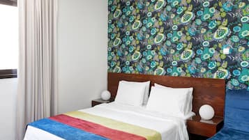 A hotel room with a bed, a blue armchair, a patterned wallpaper, and a window with curtains.