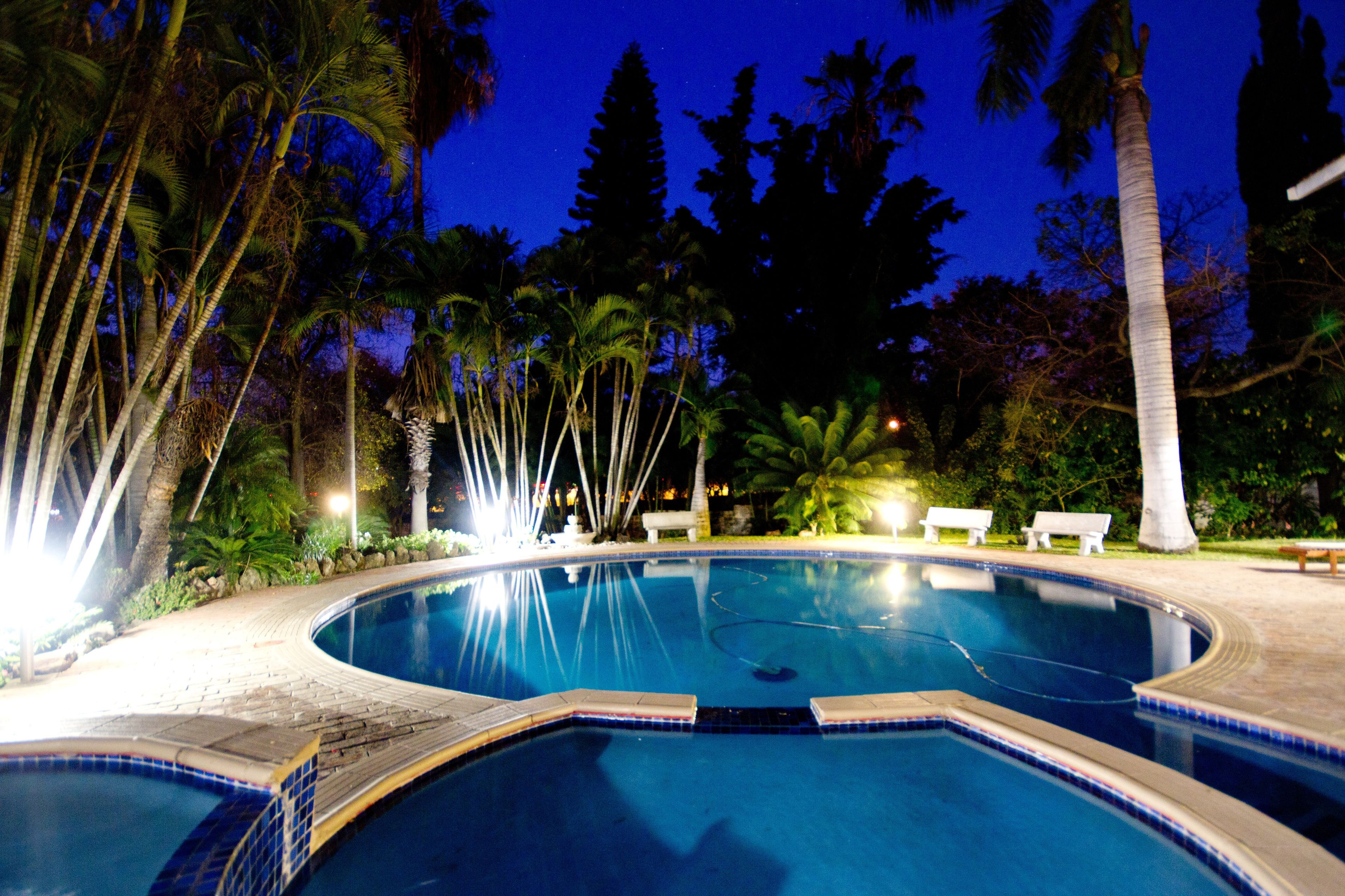 A nighttime swimming pool with a tiled edge, surrounded by palm trees and benches.