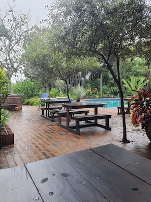 A wooden deck with picnic tables and benches, surrounded by lush greenery and a swimming pool. - Loerie Lodge (Phalaborwa)