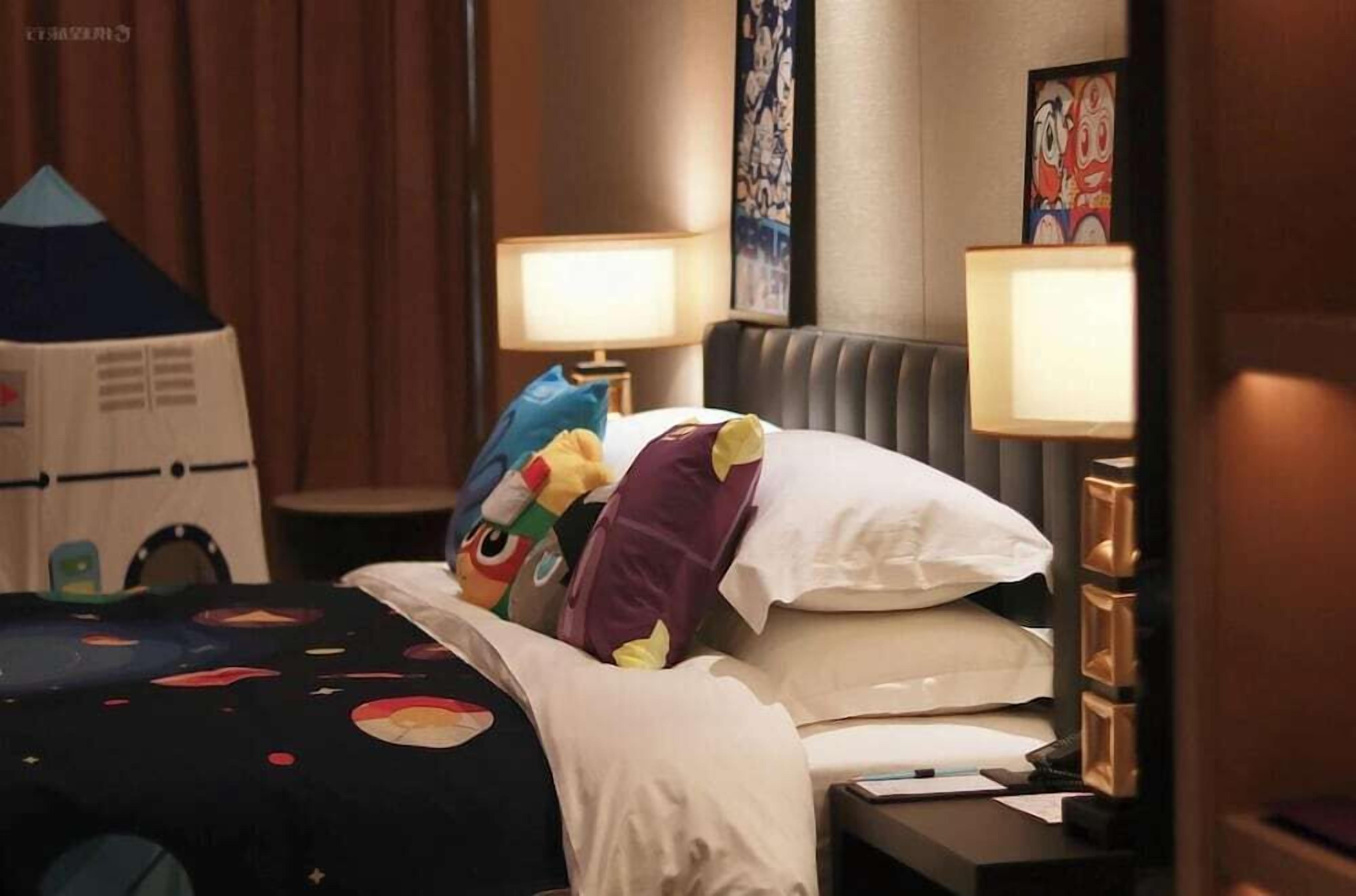 Habitación de hotel con cama, almohadas, mesita de noche, lámpara y un cartel en la pared.