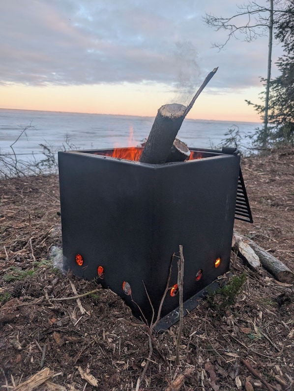 En eldstad av metall med flammor, omgiven av naturligt skräp och ett träd, nära en vattenyta.