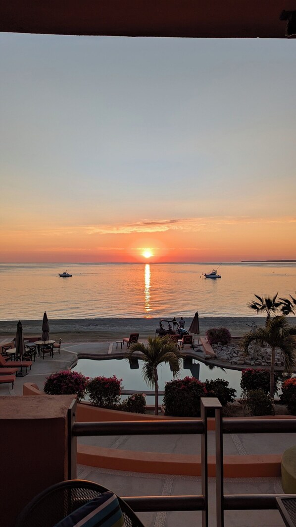 Une vue sur le coucher de soleil depuis un balcon donnant sur un espace piscine, avec des chaises longues et des parasols.