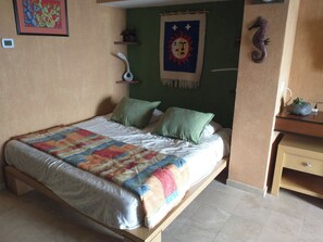 A bedroom with a bed, pillows, a wall hanging, a small shelf, and a desk with a plant.