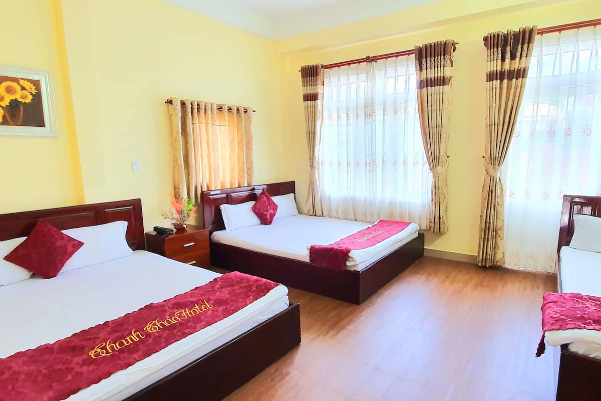 A hotel room with three beds, each with a red bedspread and a pillow. There is a nightstand between the beds, and a framed picture on the wall.