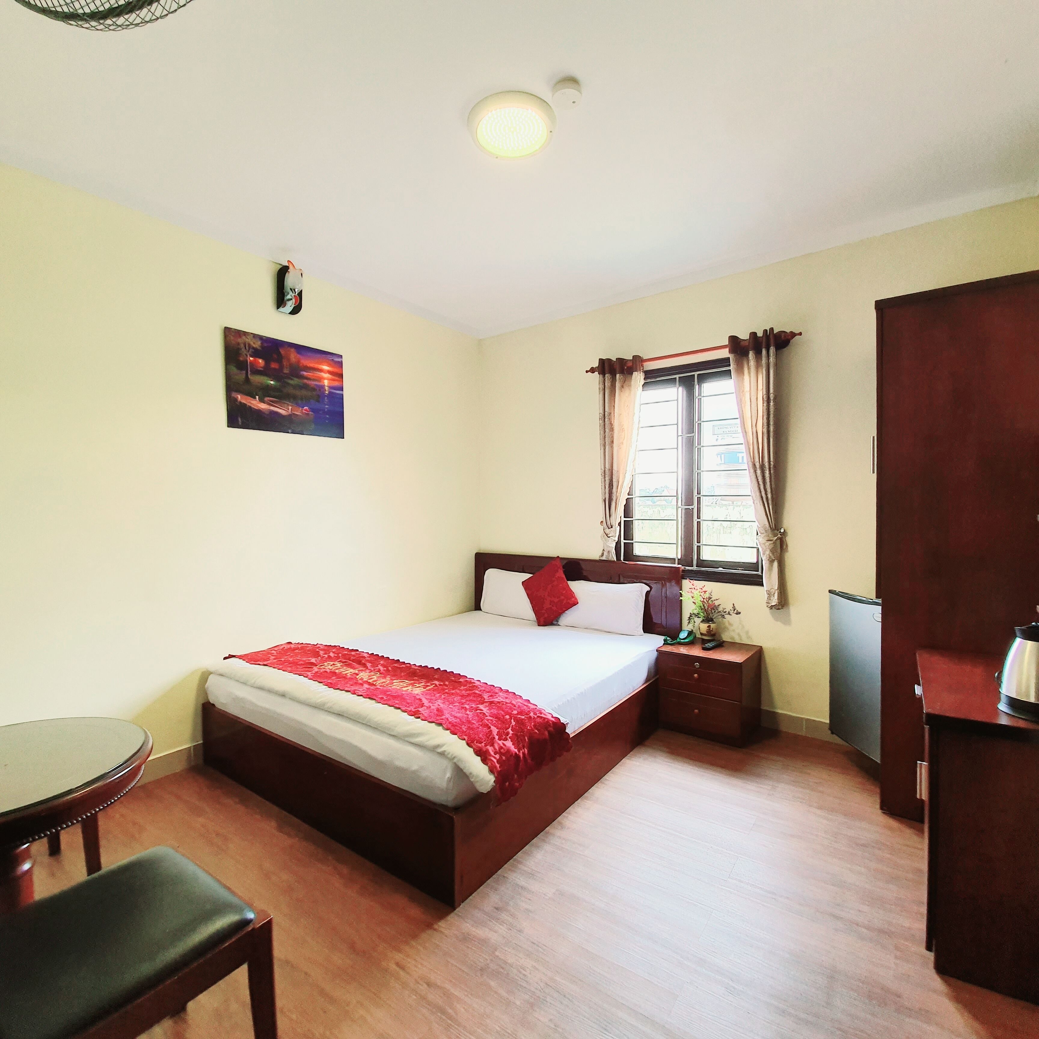A bedroom with a bed, bedside table, a small table, a chair, a window with curtains, a wall-mounted picture, and a ceiling light.