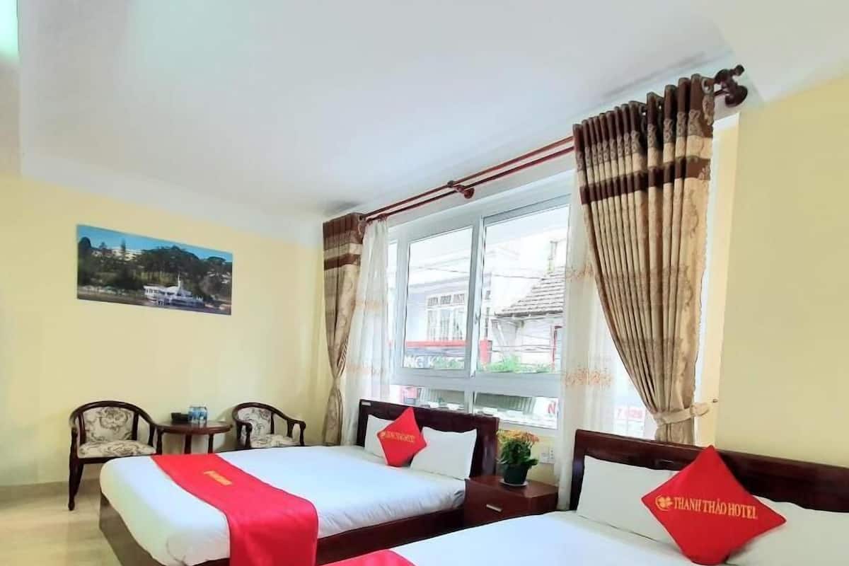 A hotel room with two beds, a window with curtains, a small table, and a chair.