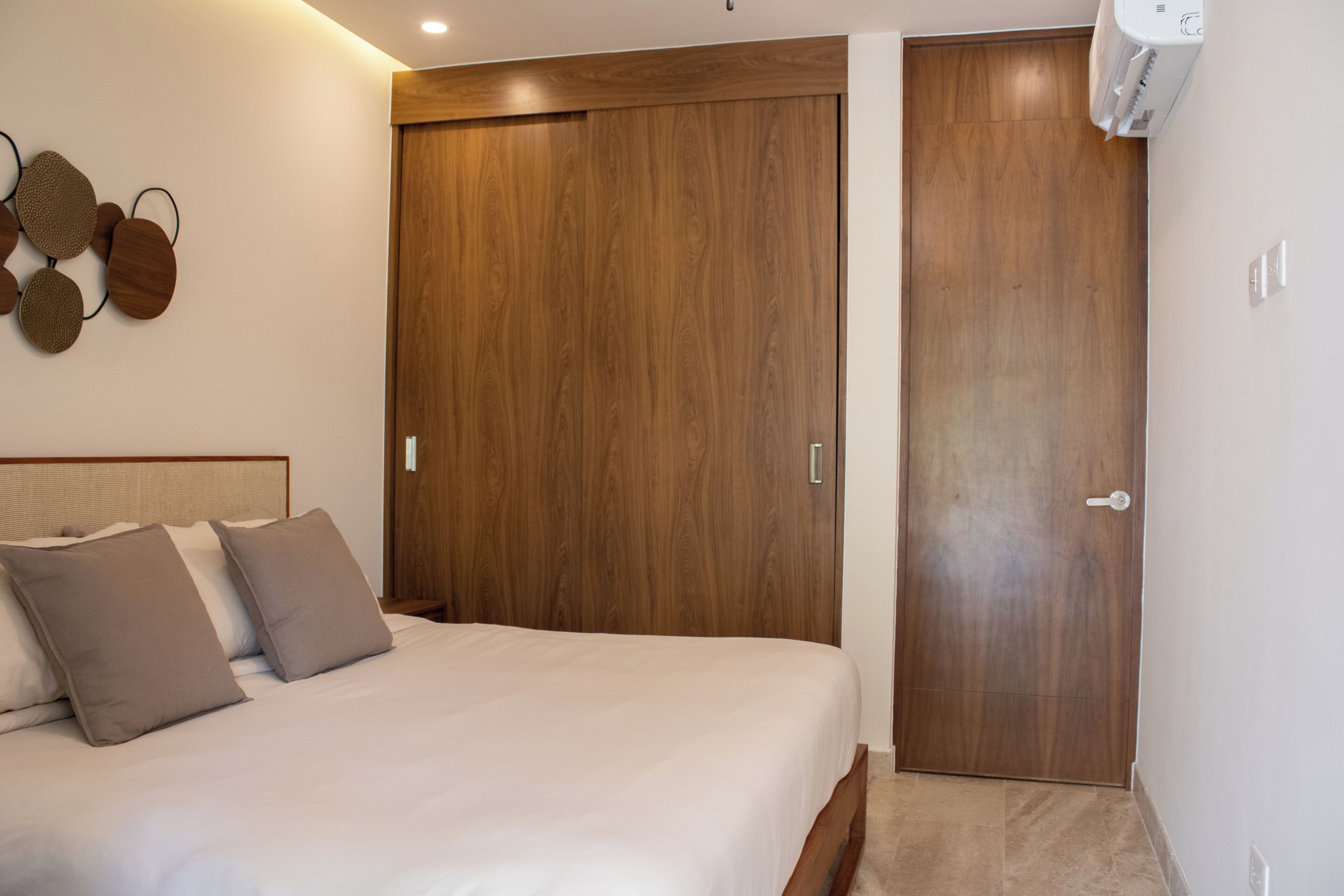 A bedroom with a bed, pillows, a wooden wardrobe, and a wall-mounted air conditioner.