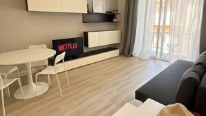 Een moderne woonkamer met een bank, een klein wit tafeltje, een televisie met een Netflix logo, een hanglamp en uitzicht door een raam met gordijnen.