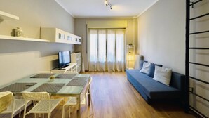 A modern living room with a sofa, a dining table with chairs, a TV, and a window with curtains.