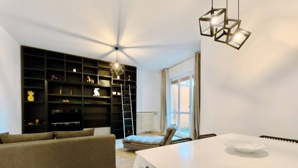 Una sala de estar moderna con un sofá, una mesa de comedor con mantel blanco, un mueble negro para estanterías y una lámpara colgante.