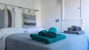 Una cama bien hecha con un edredón azul, una almohada a rayas y toallas enrolladas en una pared blanca.