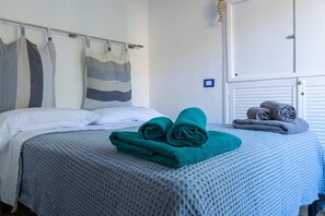 Ein ordentlich gemachtes Bett mit einer blauen Decke, einem gestreiften Kopfkissen und aufgerollten Handtüchern an einer weißen Wand.