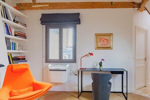 Ein Zimmer mit einem modernen Sessel, einem Schreibtisch mit Lampe, einem Bücherregal, einem Fenster mit Blick und einem gerahmten Bild an der Wand.