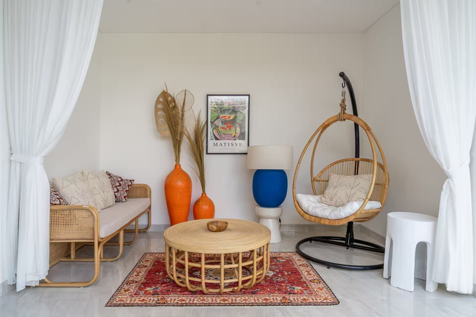 Um quarto com sofá de vime, cadeira suspensa, mesa redonda e uma obra de arte emoldurada na parede.