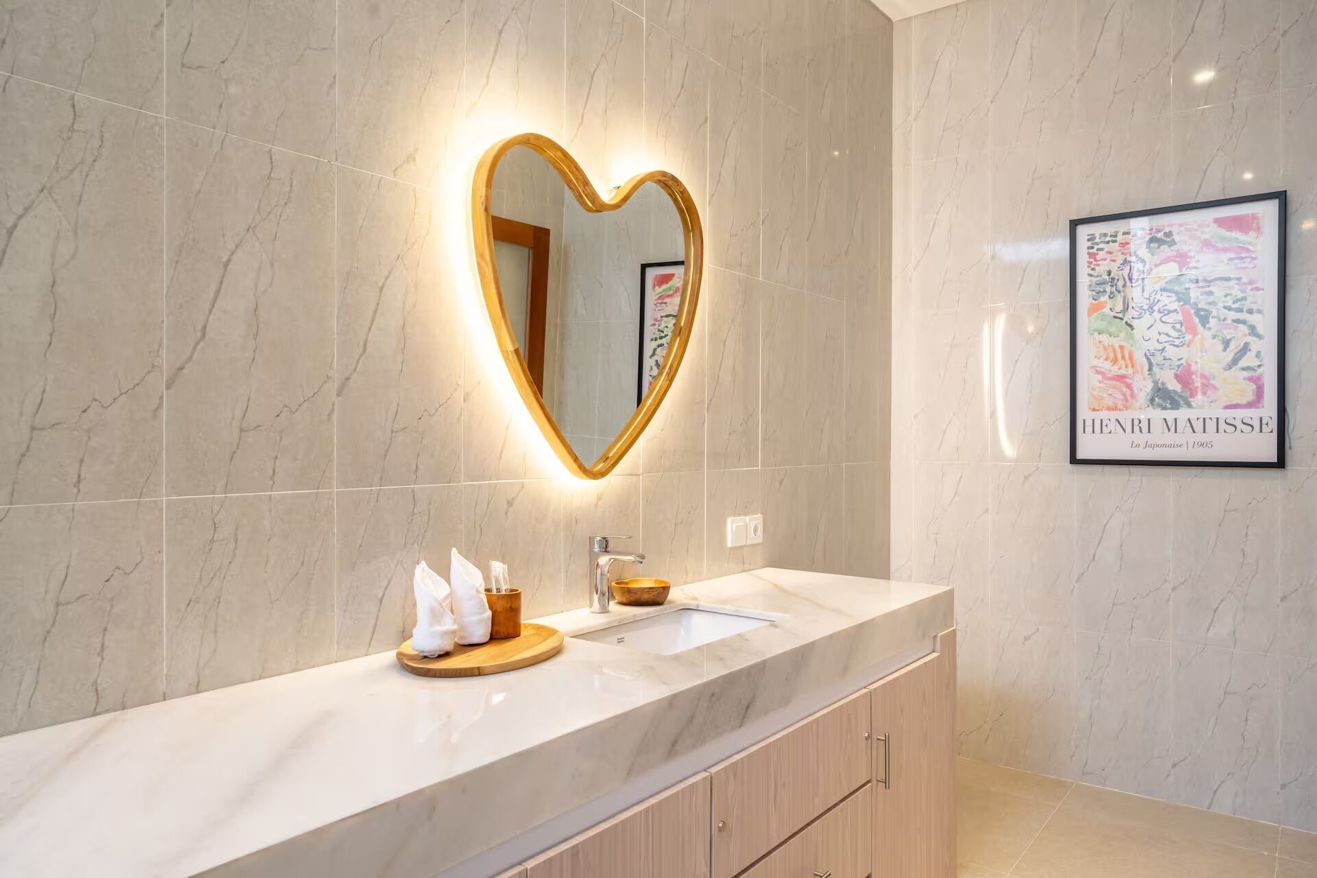 A bathroom with a heart-shaped mirror, a sink, and a framed artwork on the wall.
