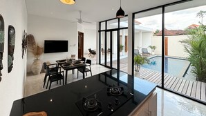 A modern kitchen with a black countertop, a built-in stove, and a dining area with a table and chairs. - Kubung Villa Bali (Kerobokan)