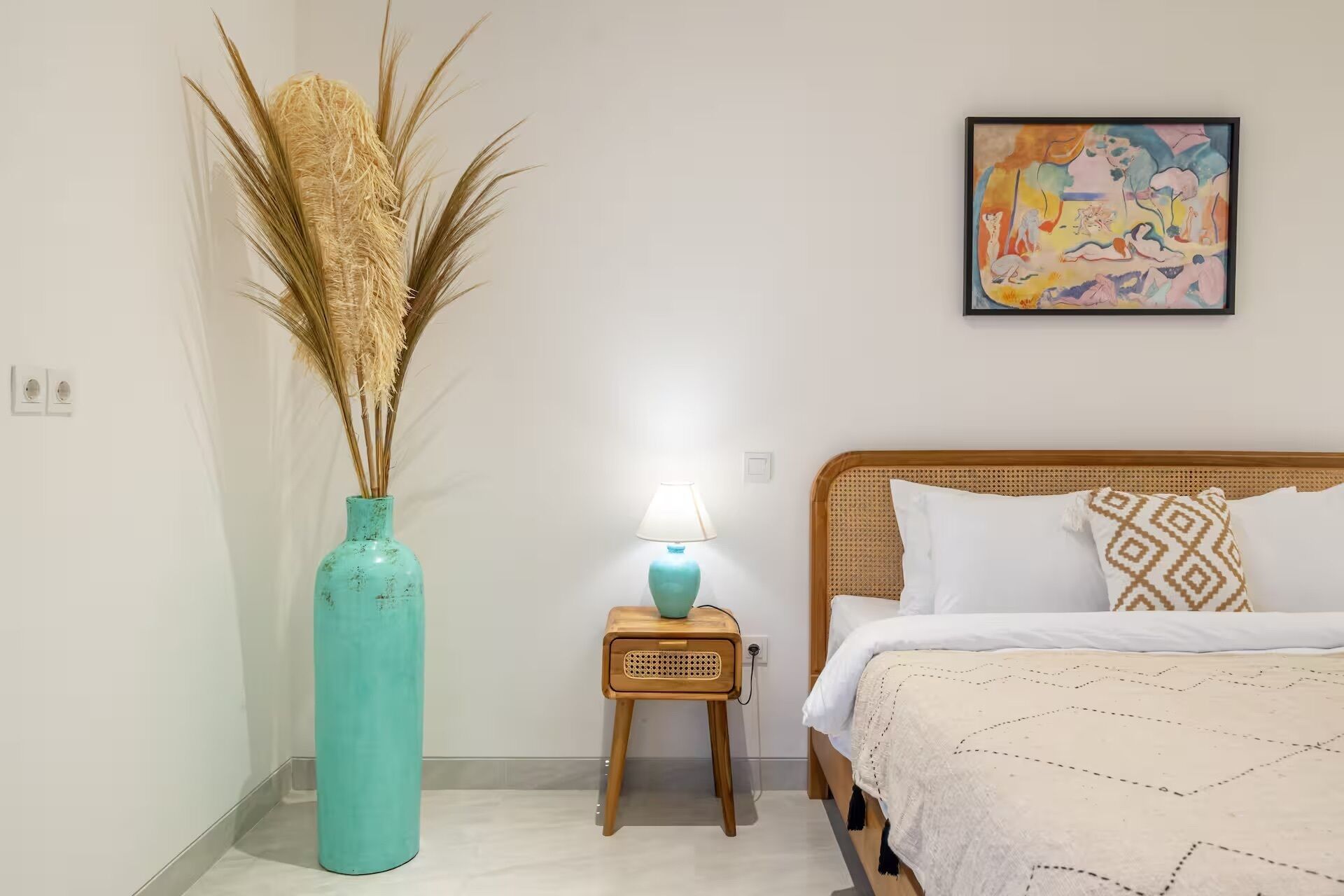 A bedroom with a bed, a bedside table with a lamp, a vase with dried plants, and a colorful painting on the wall.