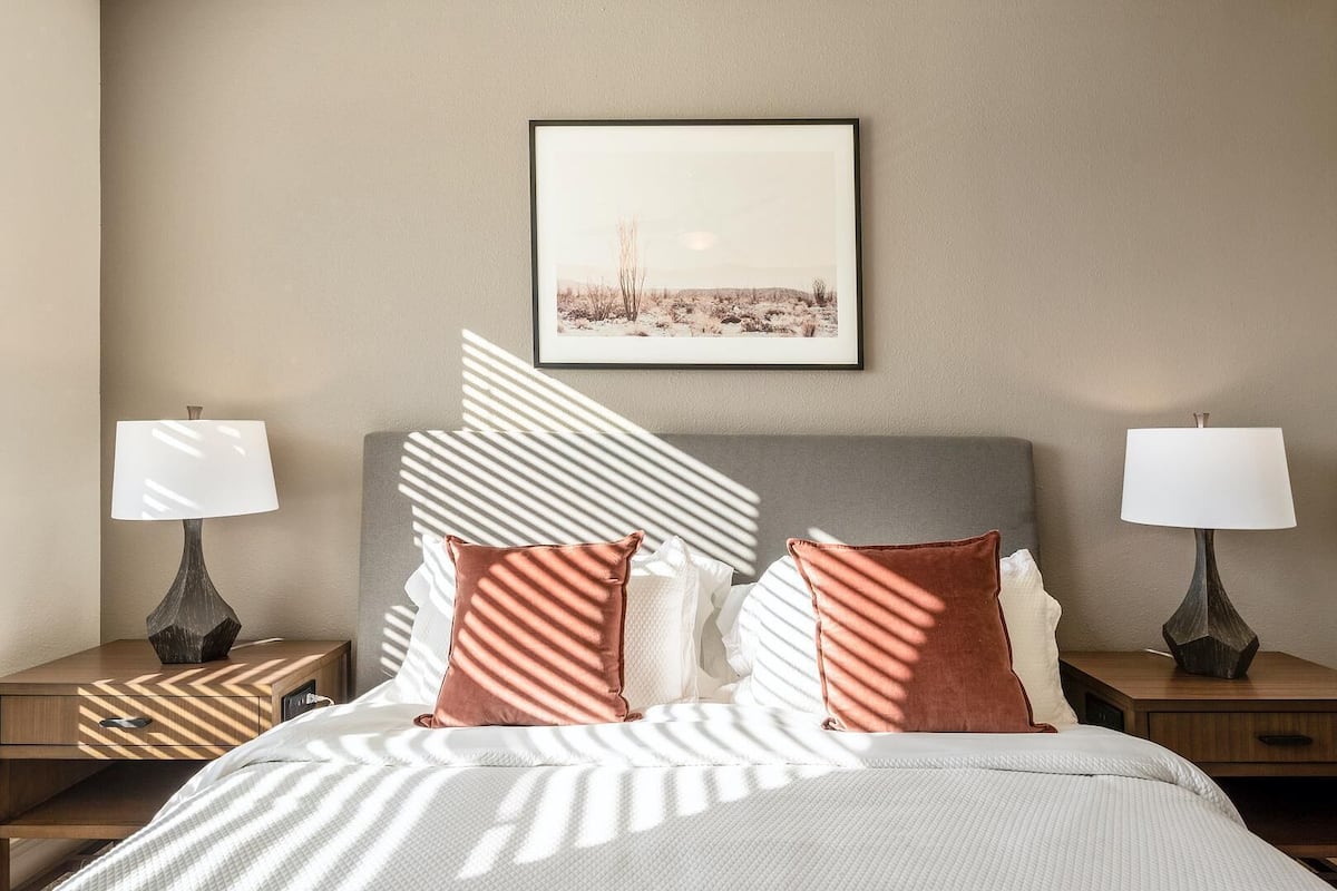Uma cama bem arrumada com manta estampada, duas luminárias de cabeceira, um quadro na parede e um padrão de sombra geométrico na parede.