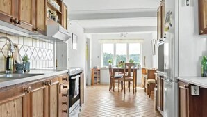 Una cocina con armarios de madera, piso de baldosas, un comedor con sillas y vistas a través de una ventana.