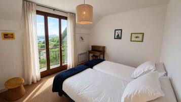 A bedroom with a large bed, a desk, a chair, a small stool, and a view of greenery through a window.