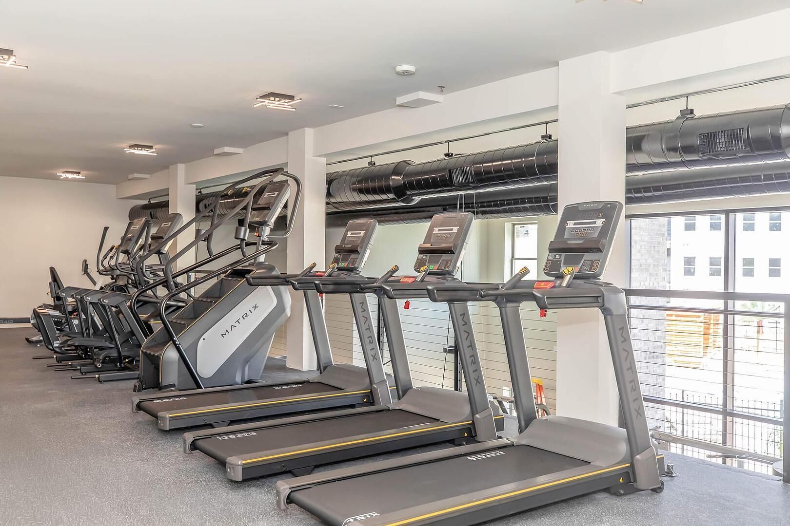 Une rangée de tapis de course dans une salle de sport, avec vue sur les bâtiments à l’extérieur.