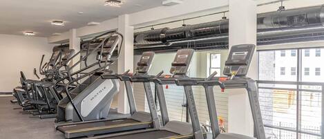Une rangée de tapis de course dans une salle de sport, avec vue sur les bâtiments à l’extérieur.
