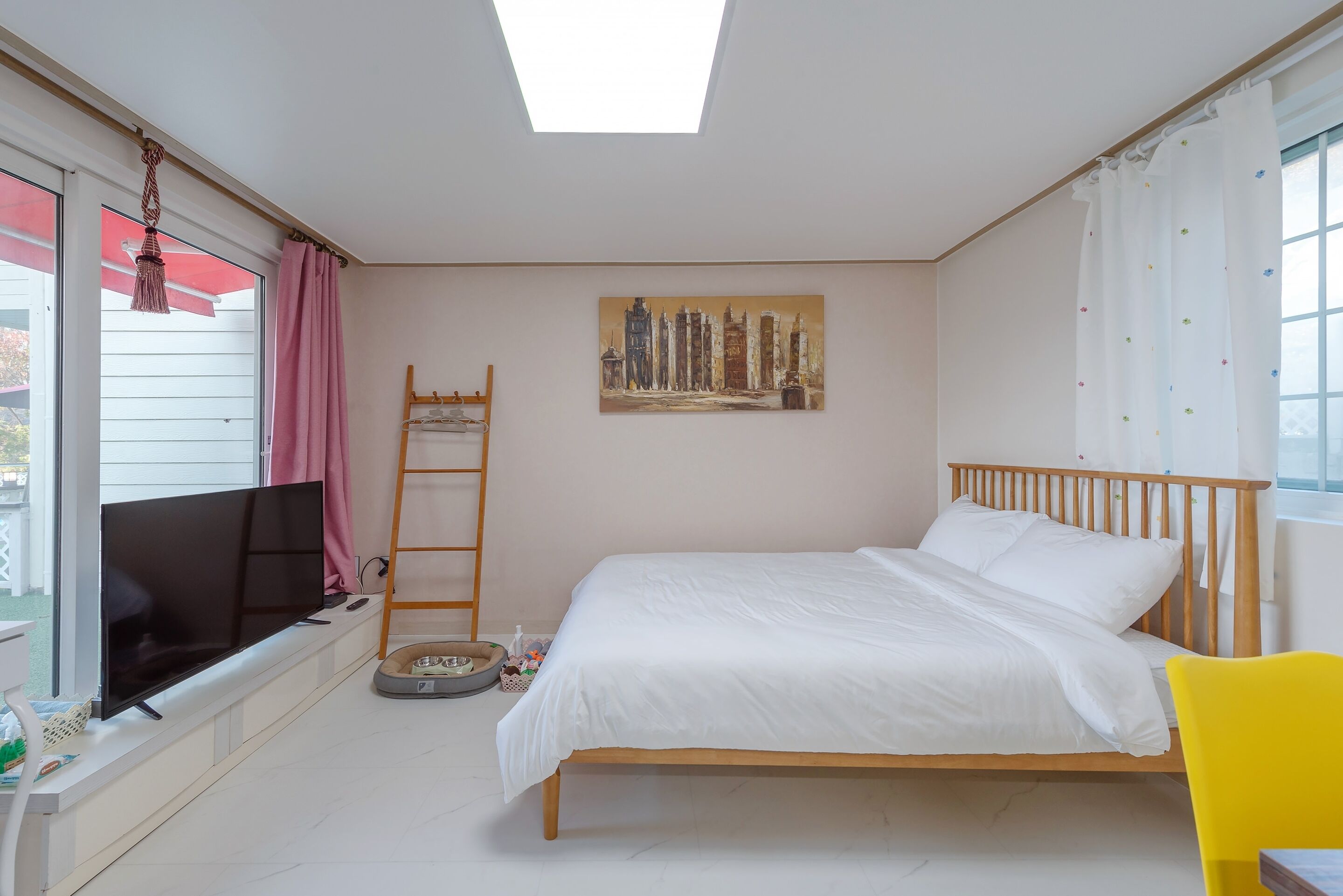 Un dormitorio con cama, televisor, un cuadro y ventana con cortinas.