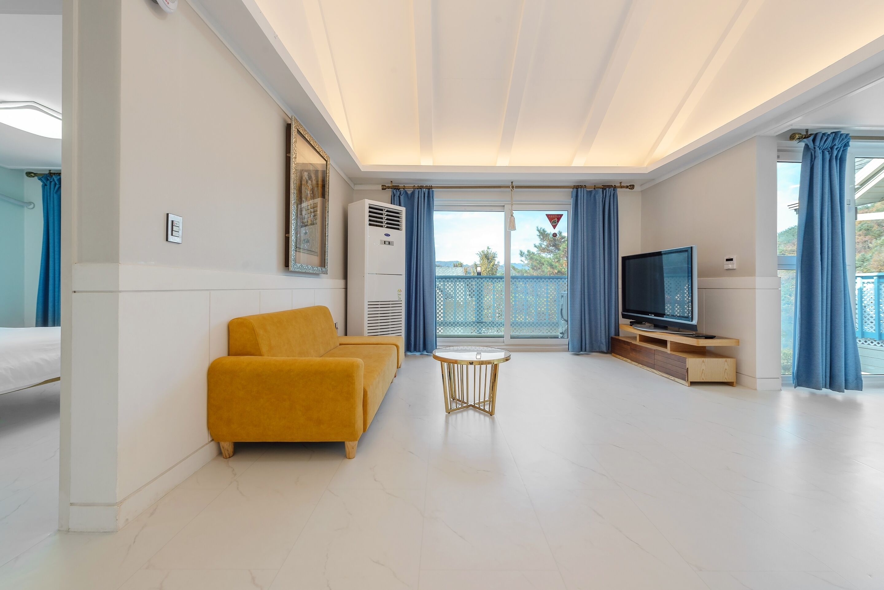 Uma sala de estar moderna com sofá, uma mesinha, televisão e varanda com cortinas azuis.