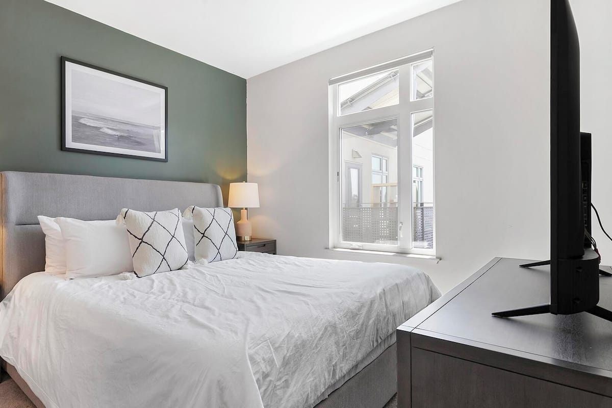 Une chambre à coucher avec un lit, deux tables de chevet avec des lampes, un tableau encadré au mur et une fenêtre donnant sur un paysage.