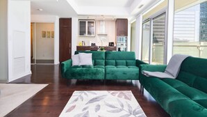 A modern living room with a green sofa, a kitchen in the background, and a large window.