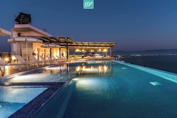 A modern infinity pool with a view of the sea, a covered outdoor seating area, and a building with a rooftop terrace. - Beachfront Lux 3BR, 2 Pools + Rooftop | Panarea 7B (Bucerías)