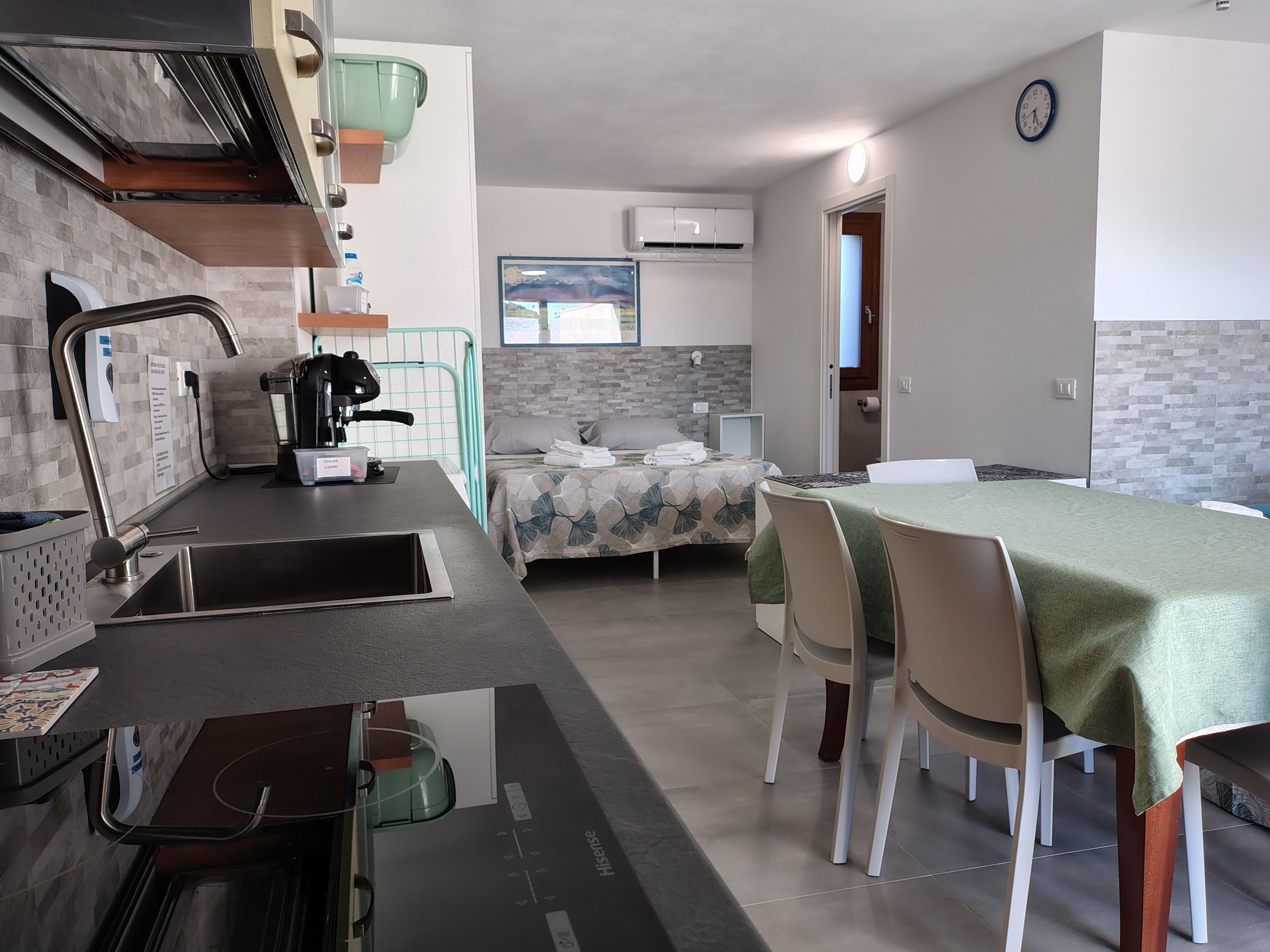 A compact hotel room with a kitchenette, a bed with a floral bedspread, a dining area with a table and chairs, and a view of the outdoors through a window.