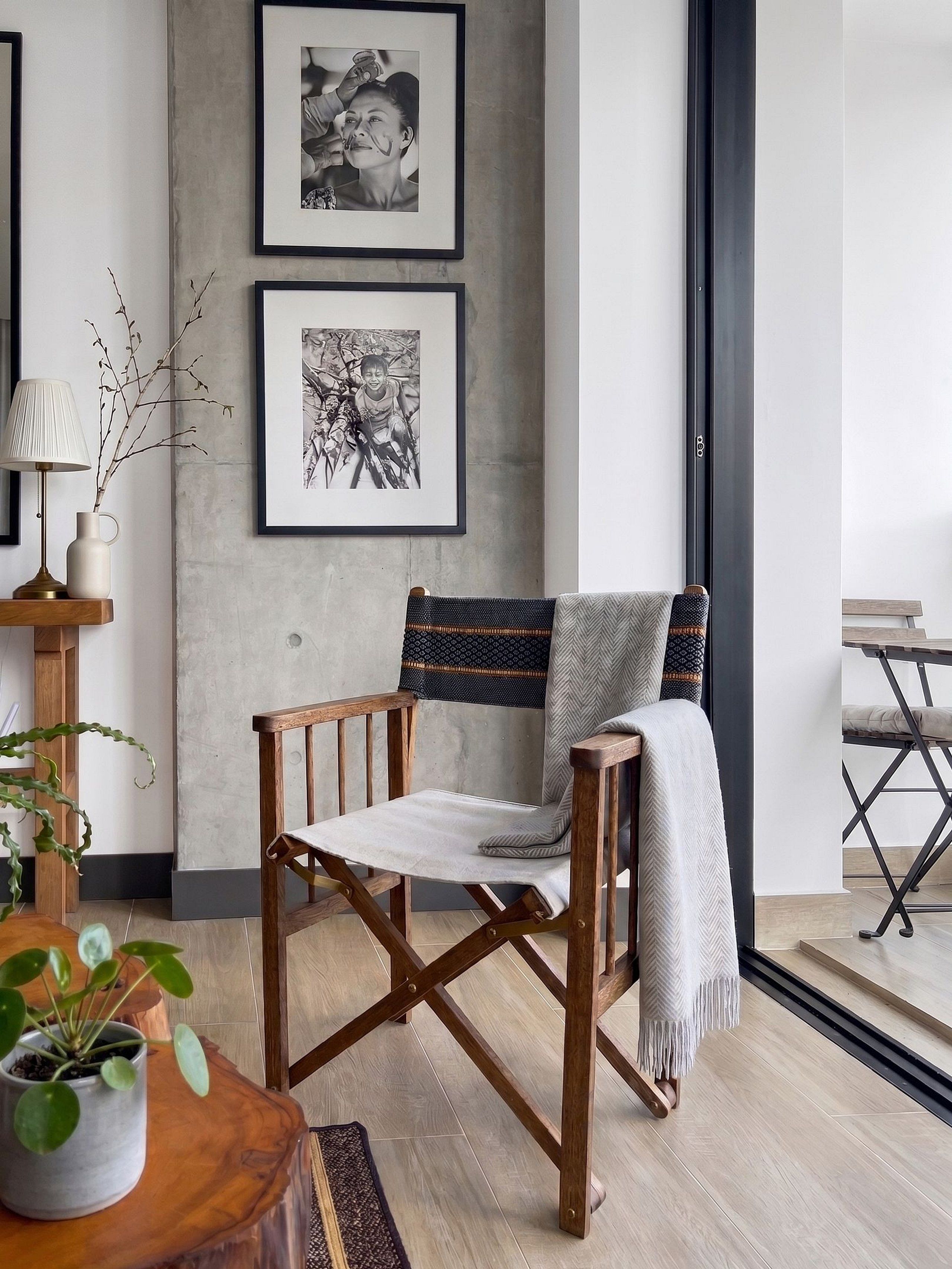 Una sala moderna con una silla de madera, una lámpara, un florero con ramas y dos fotografías en blanco y negro enmarcadas en la pared.
