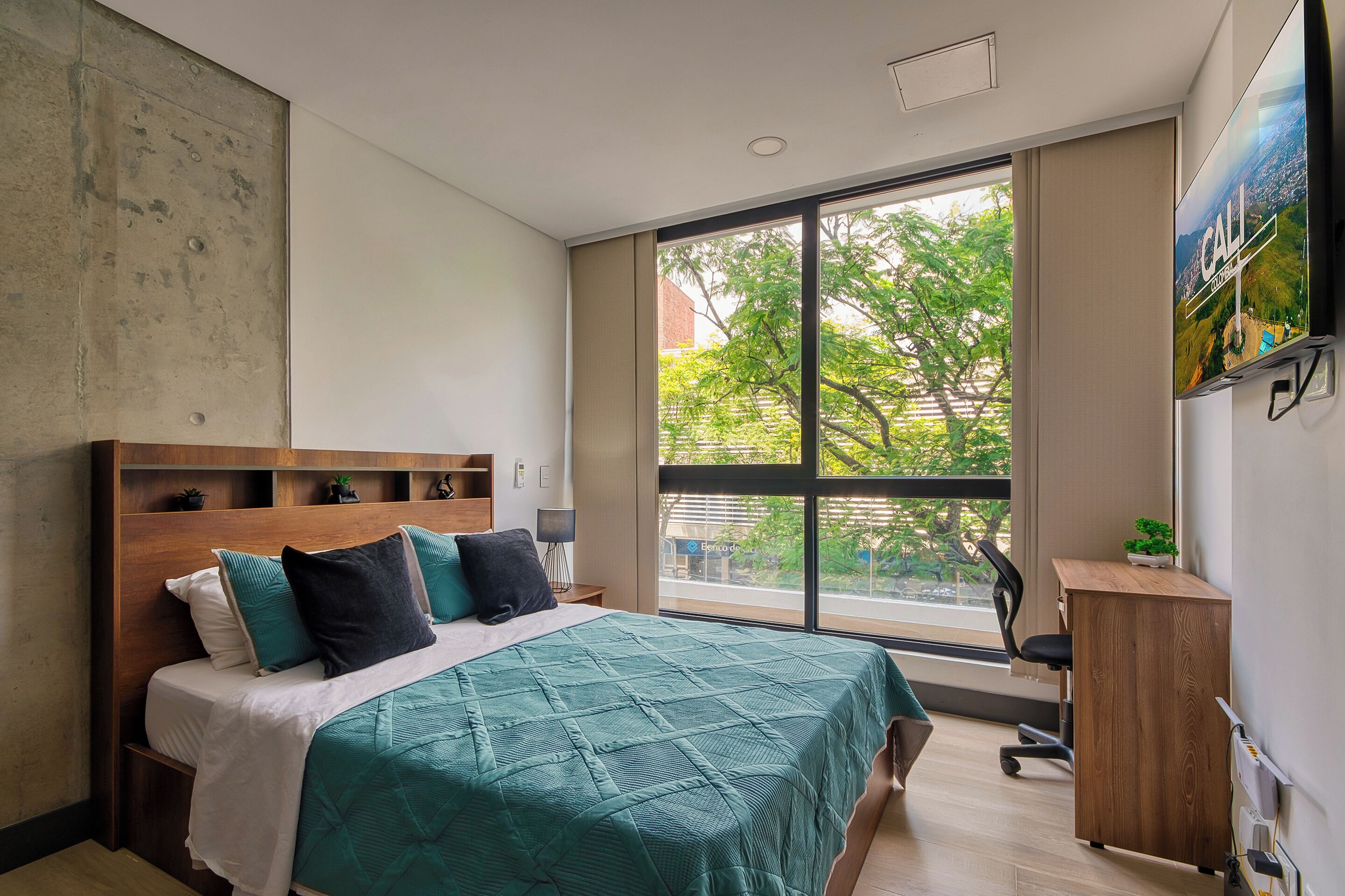 A bedroom with a large window, a bed with a green bedspread, a wooden headboard, a desk with a chair, and a TV on the wall.