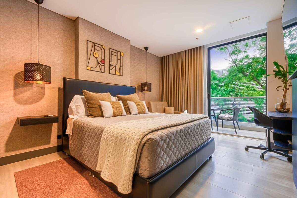 A modern bedroom with a large bed, a desk, and a view of greenery outside.