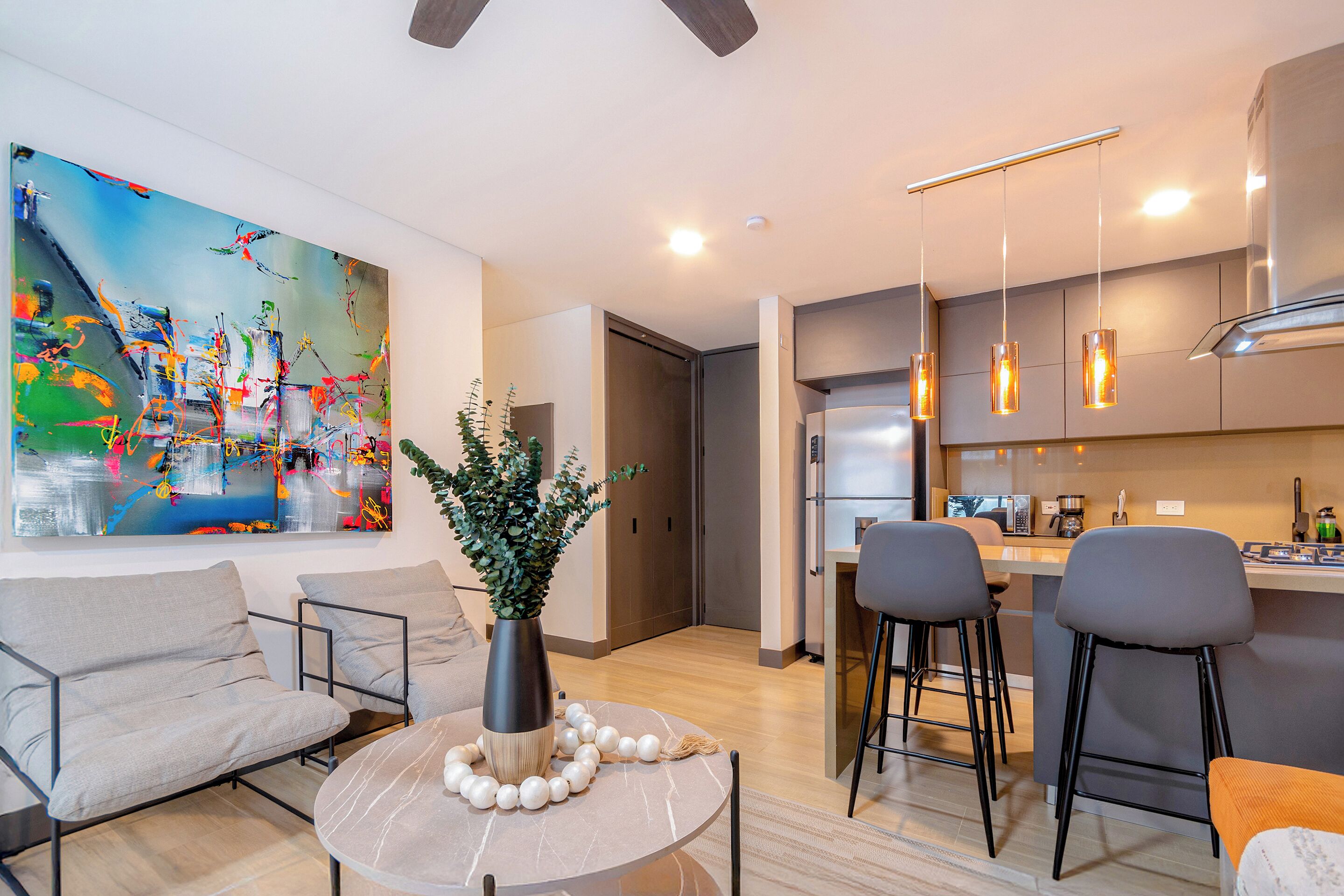 A modern living room with a round table, two sofas, a coffee table, a large abstract painting, and a kitchen area with bar stools.