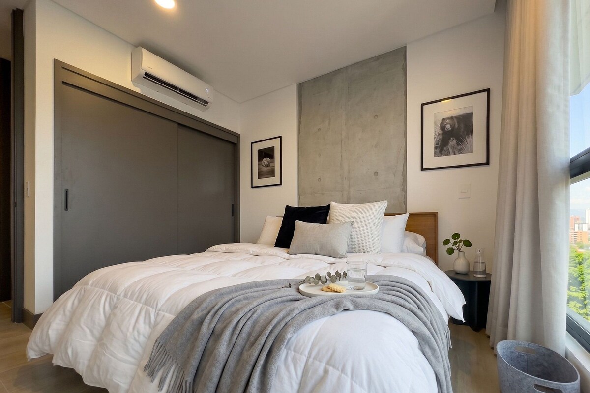 A modern bedroom with a large bed, a grey blanket, a bedside table with a vase and a glass, and a window with curtains.
