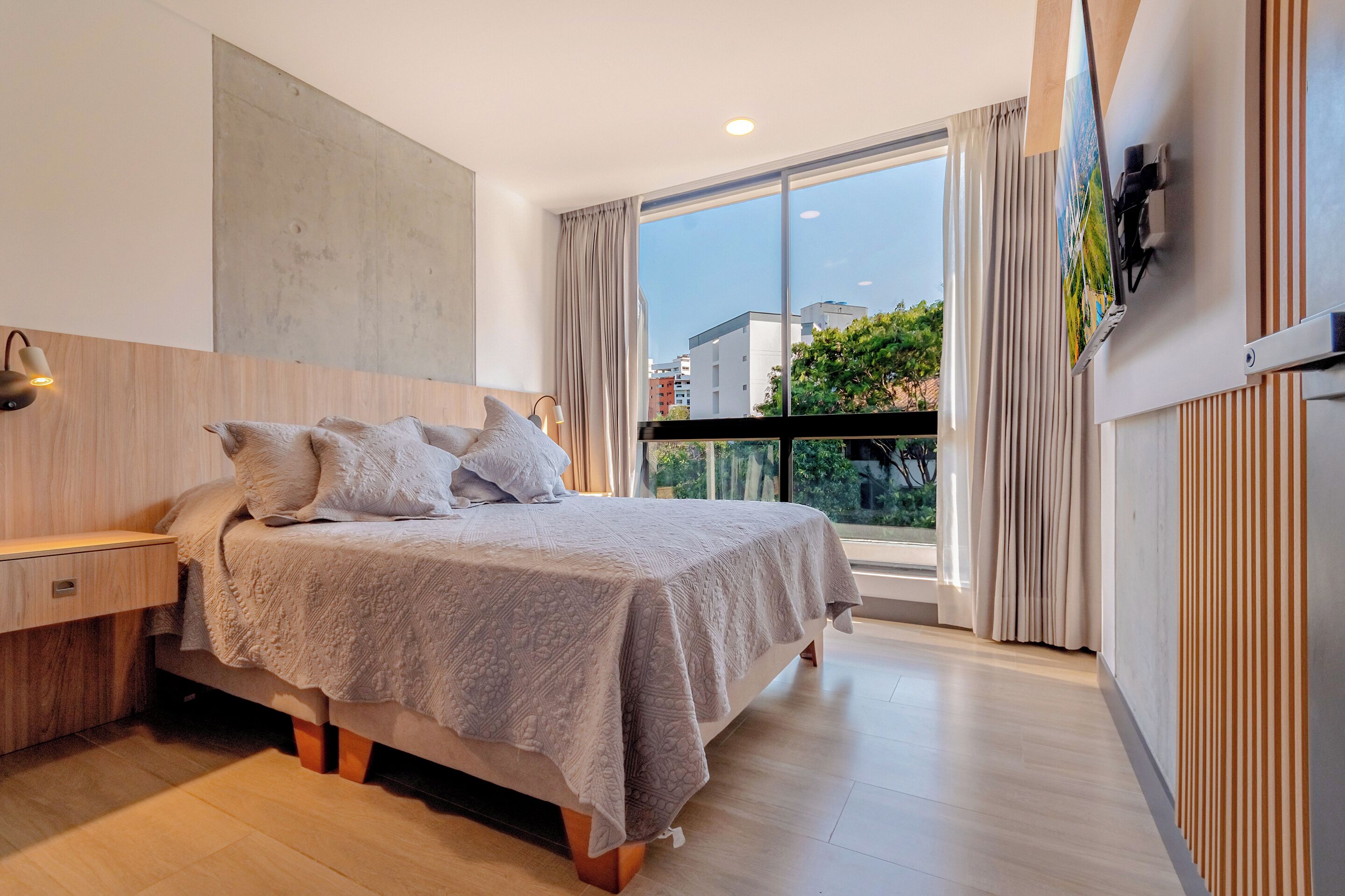 A modern bedroom with a large bed, a nightstand, a wall-mounted TV, and a balcony with curtains.